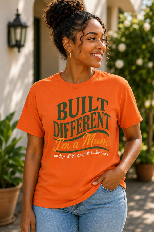 Smiling woman wearing an orange Built Different I'm a Mum graphic t-shirt with green and gold text, styled casually outdoors — Sister Style faith and heritage apparel