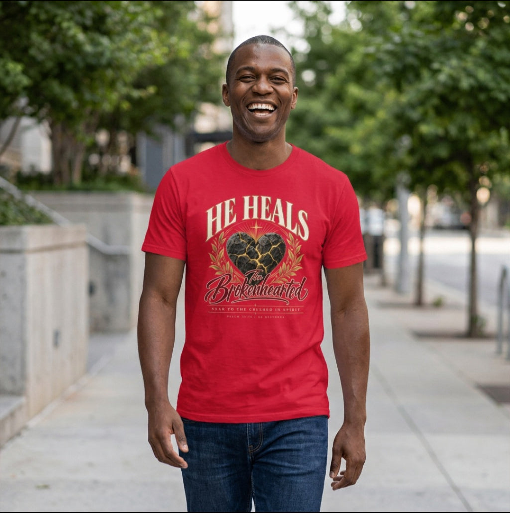 Man wearing a red Sister Style He Heals the Brokenhearted t-shirt outdoors, with a cracked heart graphic and gold-coloured leafy branches.